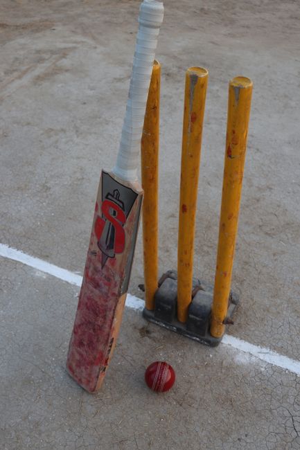 A close-up image of a bright red cricket ball with golden stitching resting on a white and navy-blue striped surface, likely a sports court or mat. The background is blurred, emphasizing the texture a